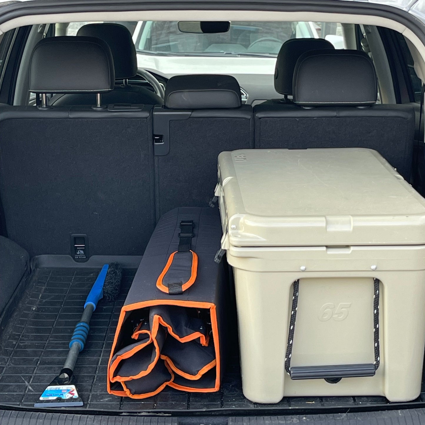 The Bug Beater 2.0 packed in an SUV trunk, positioned next to a large cooler and small windshield brush for size comparison.